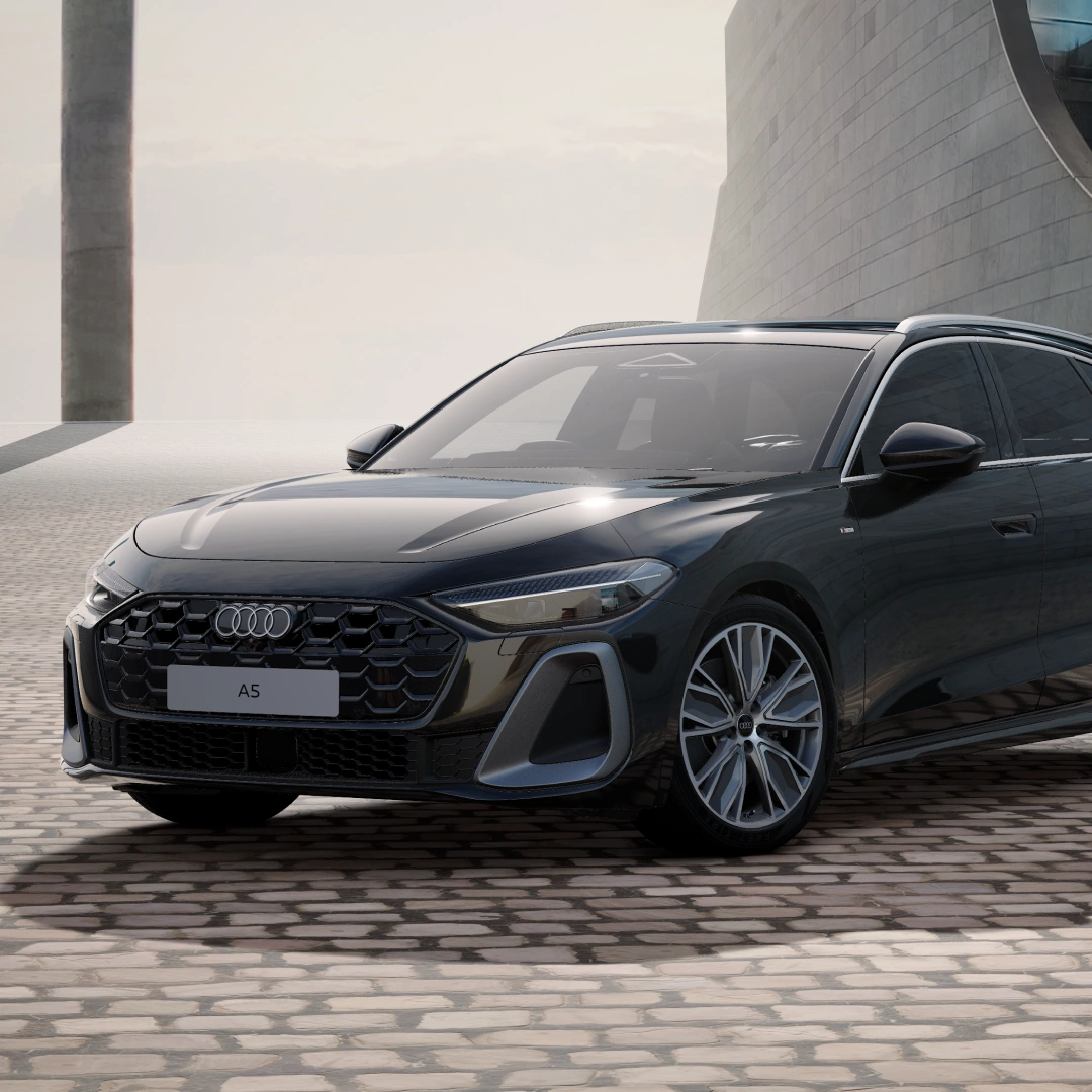 Front three-quarter view of a black Audi A5 Avant parked in a contemporary architectural setting, showcasing the Audi Singleframe grille, LED headlights, alloy wheels and sporty estate design.